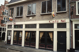 Ein Gebäude mit Fenstern und Türen, ein Schild mit der Aufschrift "Cafe Plan C in Amsterdam, Niederlande", ein Straßenschild, eine Gruppe von Bäumen und ein bewölkter Himmel.