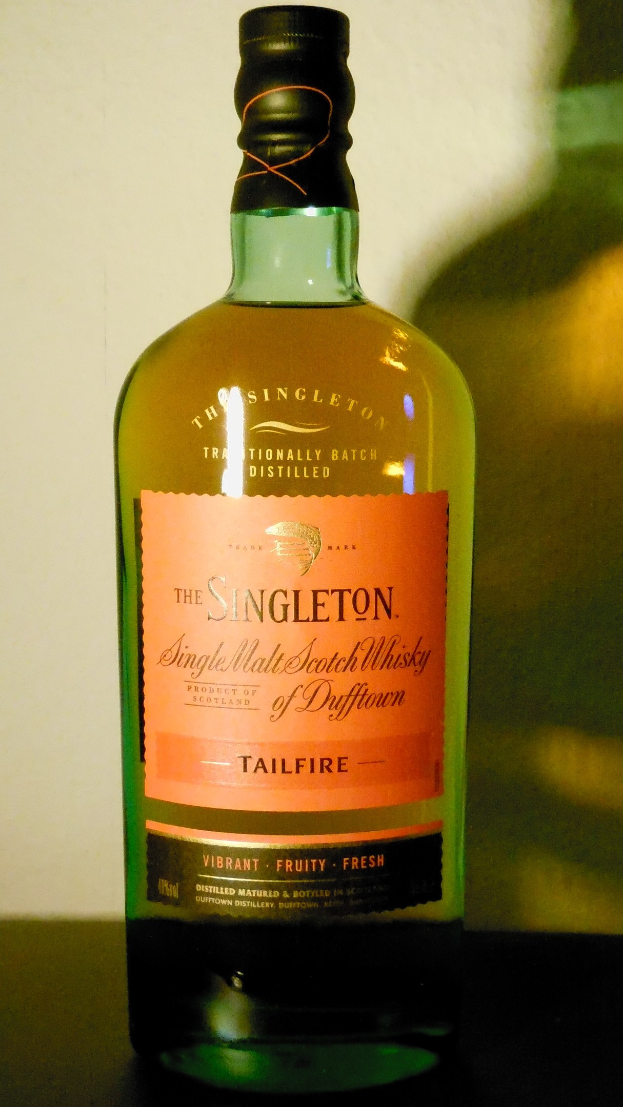 Eine tiefe bernsteinfarbene Flasche Singleton of Dufftown Tailfire Single Malt Scotch Whisky auf einem Tisch, mit einem Etikett, das eine schwarze Schrift und ein Wappen mit einem Löwen und einer Burg zeigt.