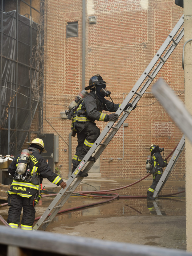Feuerwehrleute mit Helmen und Lufttanks, die eine Leiter vor einem Backsteingebäude hochklettern, mit Rohren auf dem Boden und einer Metallstange am unteren Ende, mit einem weiteren Gebäude mit Fenstern und einem Netz im Hintergrund.