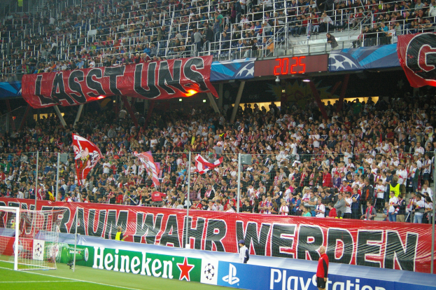 Ein Fütballspiel in einem Stadion mit einer großen Menge, saftigem Grün, einem Torpfosten links, Bodenbanner, Menschen mit Fahnen und Hintergrundgeländer, Tafeln, Lichter und andere Gegenstände.