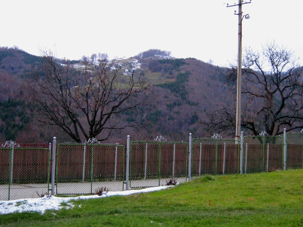 Eine grüne Landschaft mit einem Zaun, einem Pfahl und Bäumen, mit einigen Häusern im Hintergrund auf einem Hügel sichtbar
