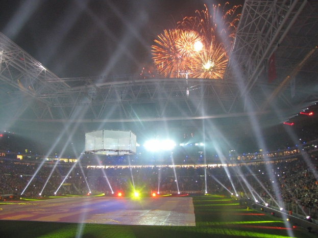 Das Olympische Stadion in London, England, bei Nacht mit heller Beleuchtung, umgeben von einer großen Menge, grünem Gras und Feuerwerk, das den Himmel erhellt.