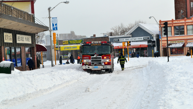 Eine Feuerwehrauto steht im Schnee vor Gebäuden, mit Geschäften und Laternen auf der linken Seite, einer Ampel in der Mitte und Schnee auf dem Boden.