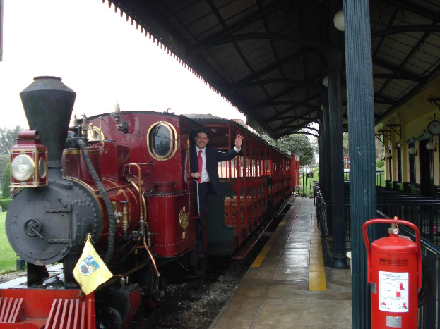 Ein alter roter Zug mit einem Mann in einem schwarzen Anzug, der an der Tür steht, lächelt und posiert, mit einer Stationshütte auf der rechten Seite und einem roten Feuerlöscher im Vordergrund.