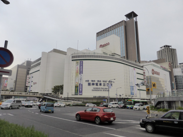Eine belebte Stra├če mit Fahrzeugen, hohen Geb├Ąuden, Stra├čenm├Âbeln, Vegetation und einem klaren Himmel, mit einem Konzertsaal mit einer Tafel im Vordergrund.