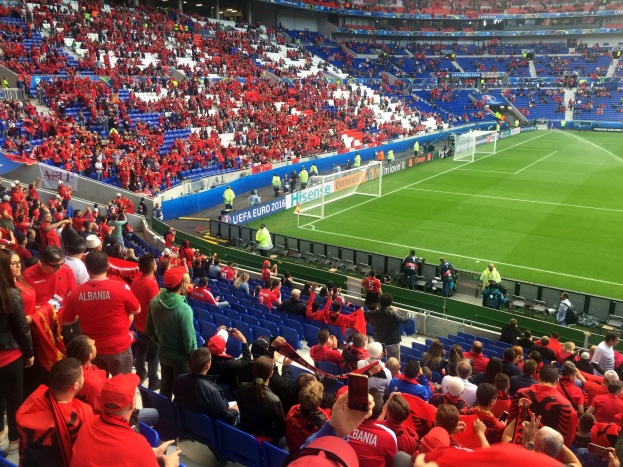 Eine große Menschenmenge in einem Stadion schaut einem Fußballspiel zu, mit Sitzplätzen, Schildern, einem Torpfosten und Deckenbeleuchtung.