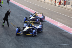 Zwei Formel-E-Autos auf einer Rennstrecke mit einem Fahne schwingenden Mann in der Nähe, einige Menschen auf der linken Seite und Geländern mit mehr Menschen im Hintergrund.