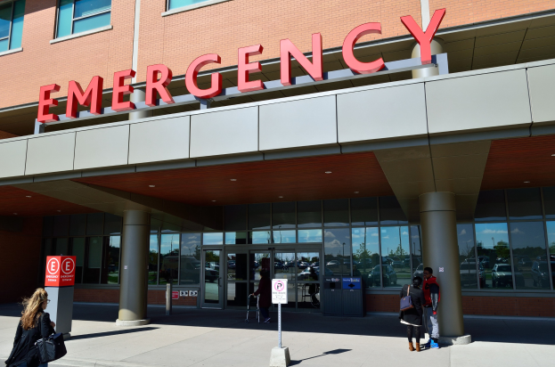 Eine Gruppe von Menschen vor einem Glaswand-Notaufnahmesgebäude mit Säulen, einem Namensschild, Schildern an Pfosten und einem Mülleimer.