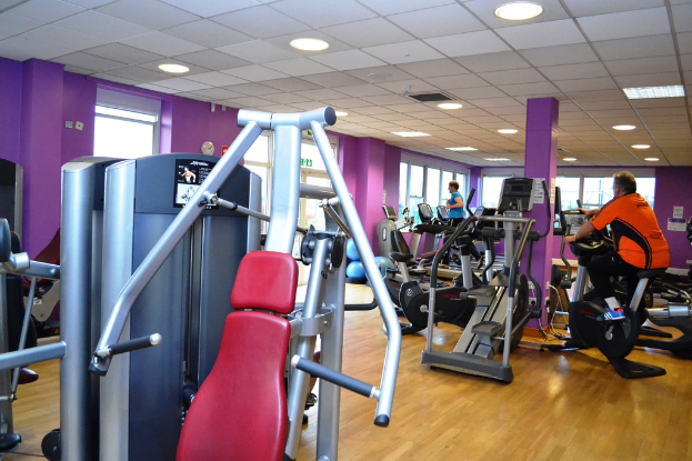 Ein Fitnessstudio mit verschiedenen Geräten und einigen Menschen beim Training, mit einer Decke und Lampen oben.