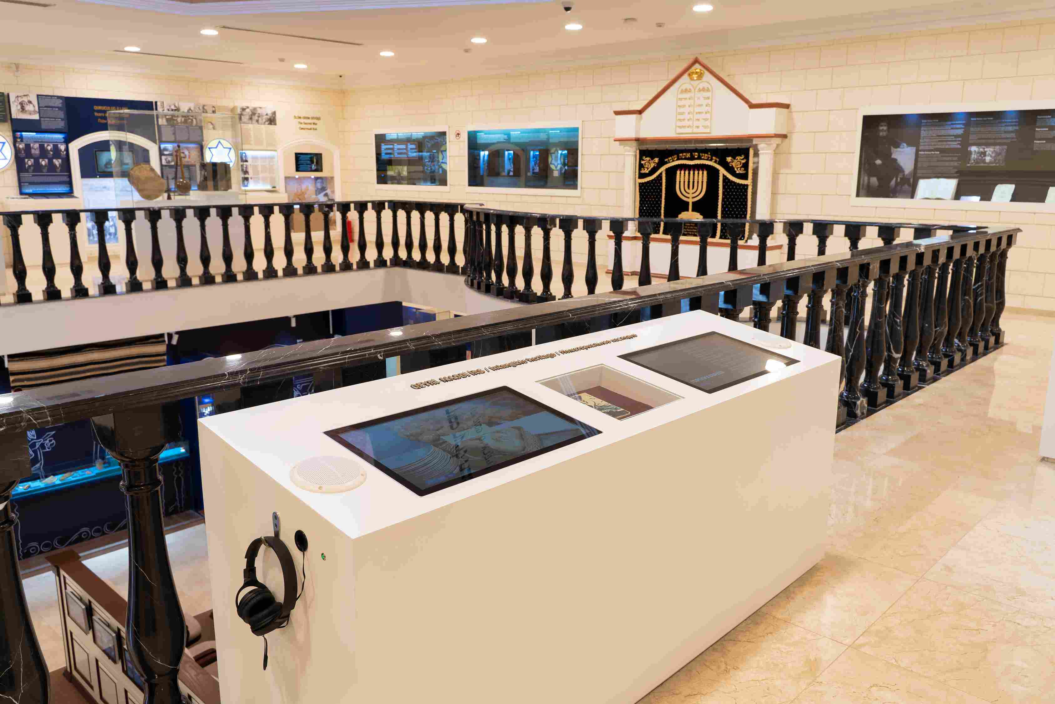 Ein Museumsraum mit verschiedenen Ausstellungsstücken, einem Tisch mit einem Headset, Geländern, wandmontierten Bildschirmen und Deckenleuchten.