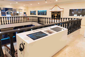 Ein Museumsraum mit verschiedenen Ausstellungsstücken, einem Tisch mit einem Headset, Geländern, wandmontierten Bildschirmen und Deckenleuchten.