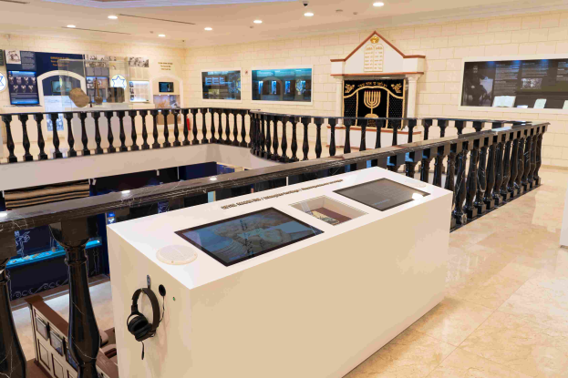 Ein Museumsraum mit verschiedenen Ausstellungsstücken, einem Tisch mit einem Headset, Geländern, wandmontierten Bildschirmen und Deckenleuchten.