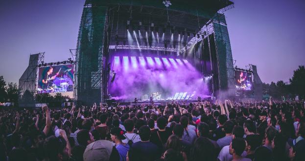 Eine große Menschenmenge versammelte sich auf einem Musikfestival, auf dem eine Bühne mit Musikern, die Instrumente spielen, beleuchtet von Lichtern und umgeben von Bildschirmen, mit einem Gebäude, Bäumen und einem klaren blauen Himmel im Hintergrund zu sehen ist.