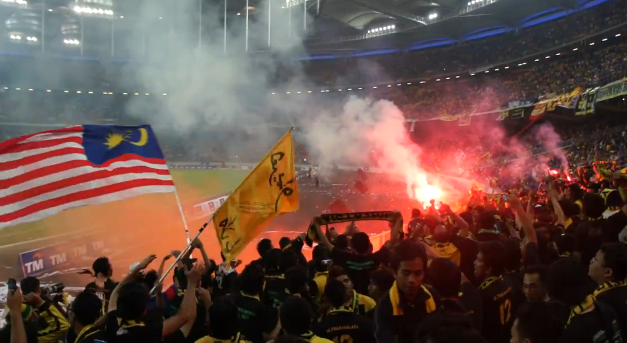 Eine Zuschauermenge in einem Stadion mit Bengalos, Fahnen und einer Kamera, mit Rauch und Schildern, die "Malaysia vs Malaysia Live Streaming" lesen, im Hintergrund.