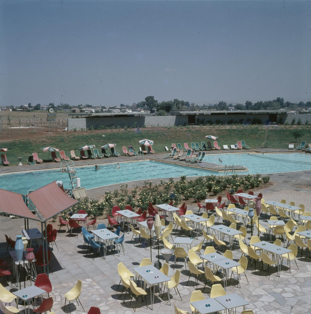 Ein Freibad mit Tischen, Stühlen und Sonnenschirmen, umgeben von Blumenpflanzen, einem Zaun, einer Wand, Bäumen, Gebäuden und unter einem bewölkten Himmel.