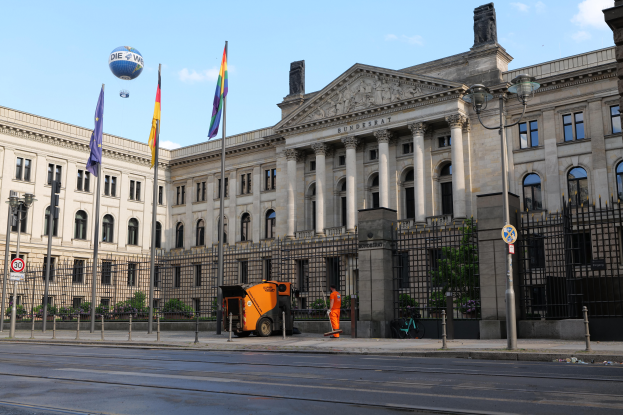 Das Reichstagsgebäude in Berlin, Deutschland, ein große Gebäude mit zahlreichen Fenstern, umgeben von Straßenmasten, Fahrzeugen, Menschen und Bäumen, unter einem bewölktem Himmel.