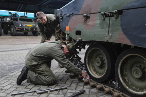 Zwei Männer in militärischer Uniform arbeiten an einem Militärfahrzeug in einer Garage, umgeben von Werkzeugen, mit anderen Militärfahrzeugen und einem Schuppen im Hintergrund.