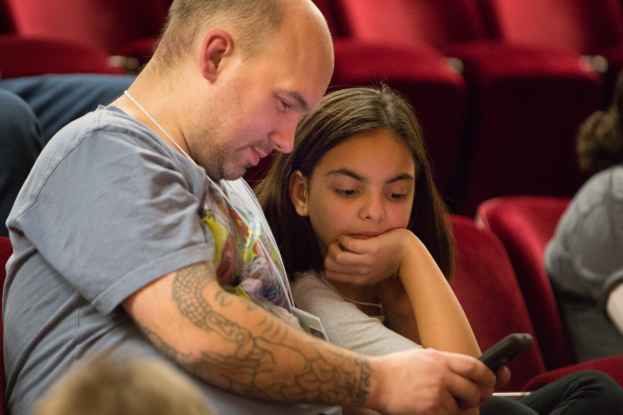 Ein Mann und ein junges Mädchen sitzen in einem Kino, beide schauen auf ein Handy, wobei der Mann es hält und das Mädchen es aufmerksam betrachtet.