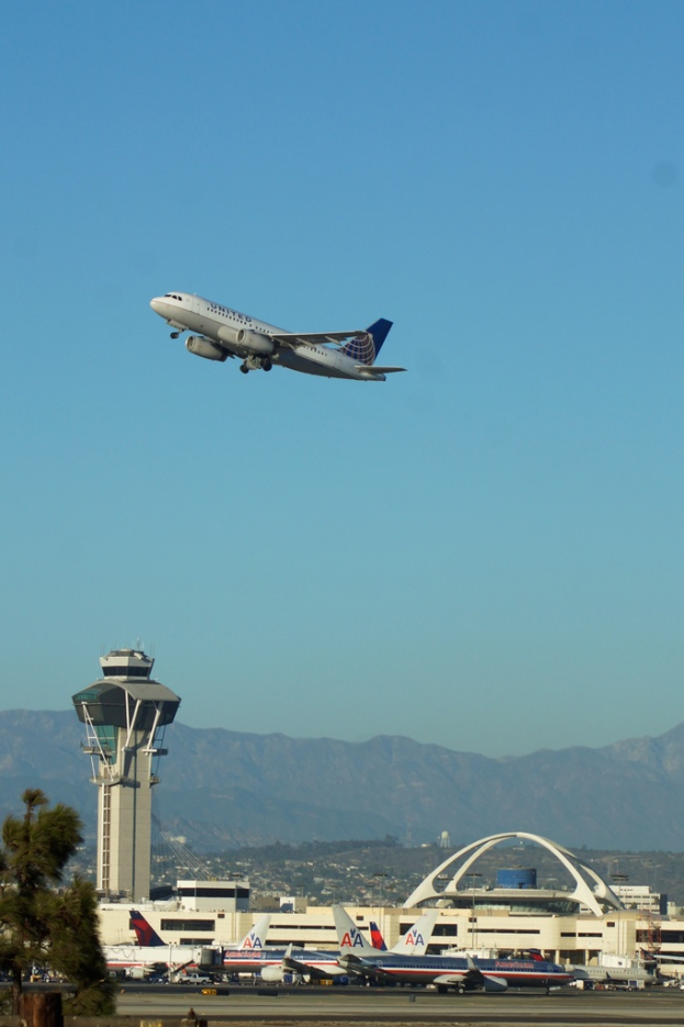 Ein Flughafenblick mit einem weißen Kontrollturm, geparkten Flugzeugen, entfernten Gebäuden und einem Flugzeug am Himmel.