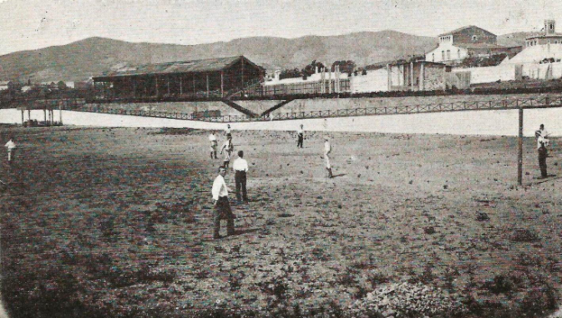 Ein Schwarz-Weiß-Foto einer Gruppe von Menschen, die in den frühen 1900er Jahren Cricket am Strand spielen, mit Gebäuden, Bäumen, Hügeln und einer Brücke im Hintergrund und Text unten.