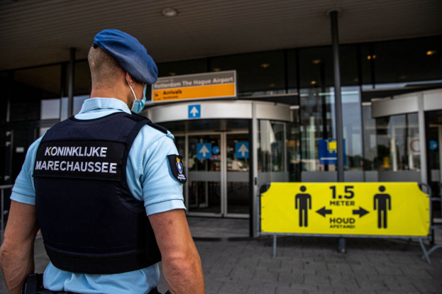 Ein Polizist in blauer Uniform und Mütze steht vor einem gläsernen Gebäude mit einem gelben Schild auf der rechten Seite und zusätzlichen Schildern im Hintergrund.