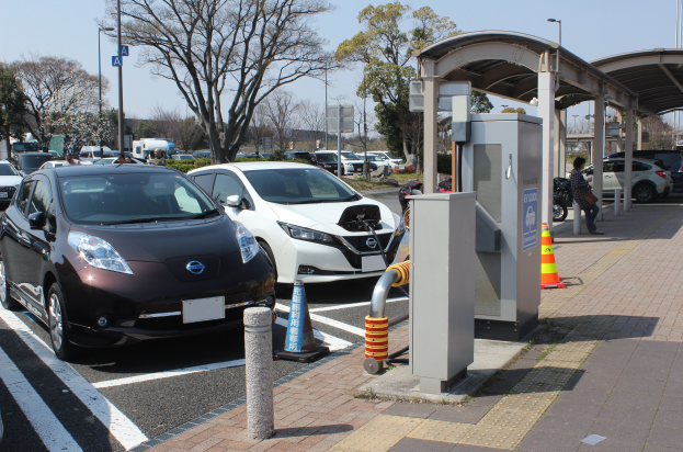 Elektroauto-Ladestation in Japan mit Autos, Verkehrskegeln, einer Person auf dem Gehweg, einem Schuppen, Masten, Lichtern, Schildern, Bäumen, Pflanzen und einem sichtbaren Himmel.