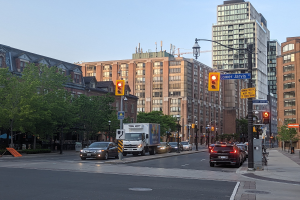 Eine belebte Stadtstraße mit einem Lastwagen, einem Auto und einem SUV im Verkehr, umgeben von Laternenmasten, Ampeln, Texttafeln, Bäumen und Gebäuden unter einem Himmel.