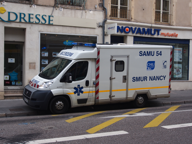 Ein Rettungswagen steht auf der Straßenseite vor einem Krankenhausgebäude mit Fenstern, Geländern, Texttafeln, Plakaten und anderen Gegenständen.