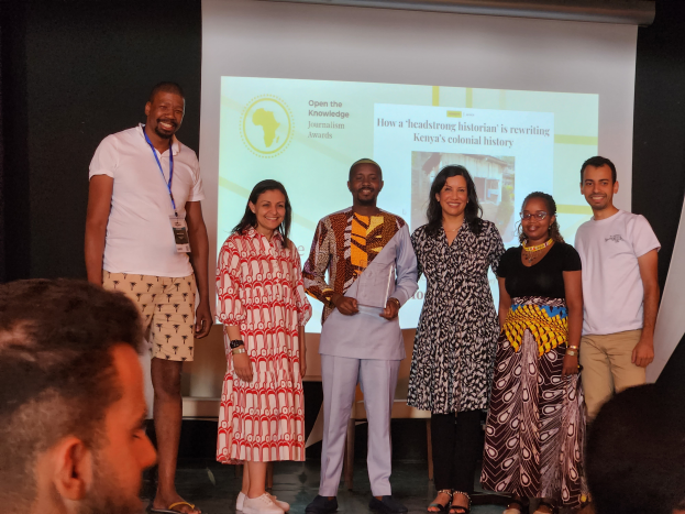 Eine Gruppe von Menschen auf einer Bühne, die lächeln und für ein Foto posieren, mit einem Publikum darunter und einem Bildschirm im Hintergrund, der 'Open the Knowledge Journalism Awards 2019' anzeigt.