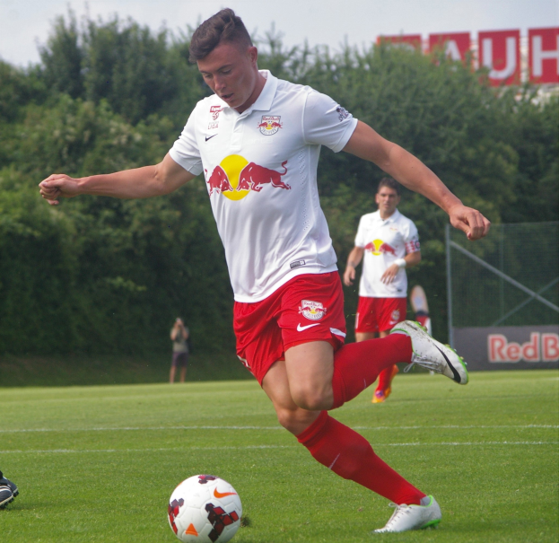 Ein Mann in einem Fußballtrikot kickt einen Ball auf einem grünen Feld mit Bäumen und einem klaren blauen Himmel im Hintergrund, mit einem Schild mit der Aufschrift "RB Leipzig".