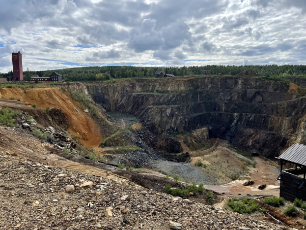 Großer Tagebau auf einem Schotterfeld umgeben von Bäumen, Gebäuden und einer Hütte unter einem bewölkten Himmel, mit verstreuten Steinen und Pflanzen auf dem Boden.