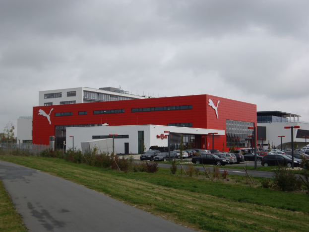Die Puma-Fabrik in Deutschland mit Industriegebäuden, Fahrzeugen auf der Straße, Straßenmasten, Straßenlaternen, Bäumen, Pflanzen, Gras und einem bewölkten Himmel.