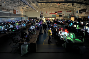 Ein großer Raum mit vielen Menschen, die Videospiele spielen, sitzend und stehend, umgeben von Monitoren, Tastaturen, Mäusen und Kabeln auf Tischen, mit markierten Platten im Hintergrund und einer sichtbaren Decke.