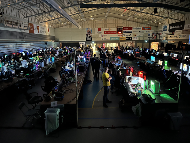 Ein großer Raum mit vielen Menschen, die Videospiele spielen, sitzend und stehend, umgeben von Monitoren, Tastaturen, Mäusen und Kabeln auf Tischen, mit markierten Platten im Hintergrund und einer sichtbaren Decke.