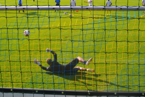 Ein Fußballspieler taucht ab, um vor einem Tor einen Ball zu halten, mit einem Netz im Vordergrund und einigen Menschen im Hintergrund, während mehrere Fußballschuhe auf dem Boden verstreut sind.