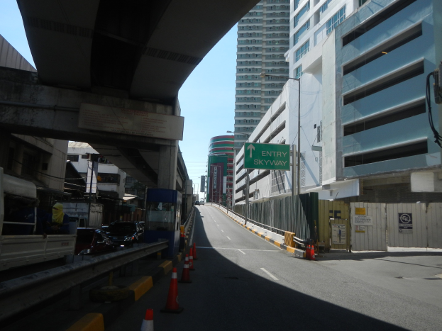 Stadtstraße mit Verkehrskegeln, Fahrzeugen, einer Brücke auf der linken Seite, Gebäuden mit Schildern, Laternenmästen und einem Himmel im Hintergrund.