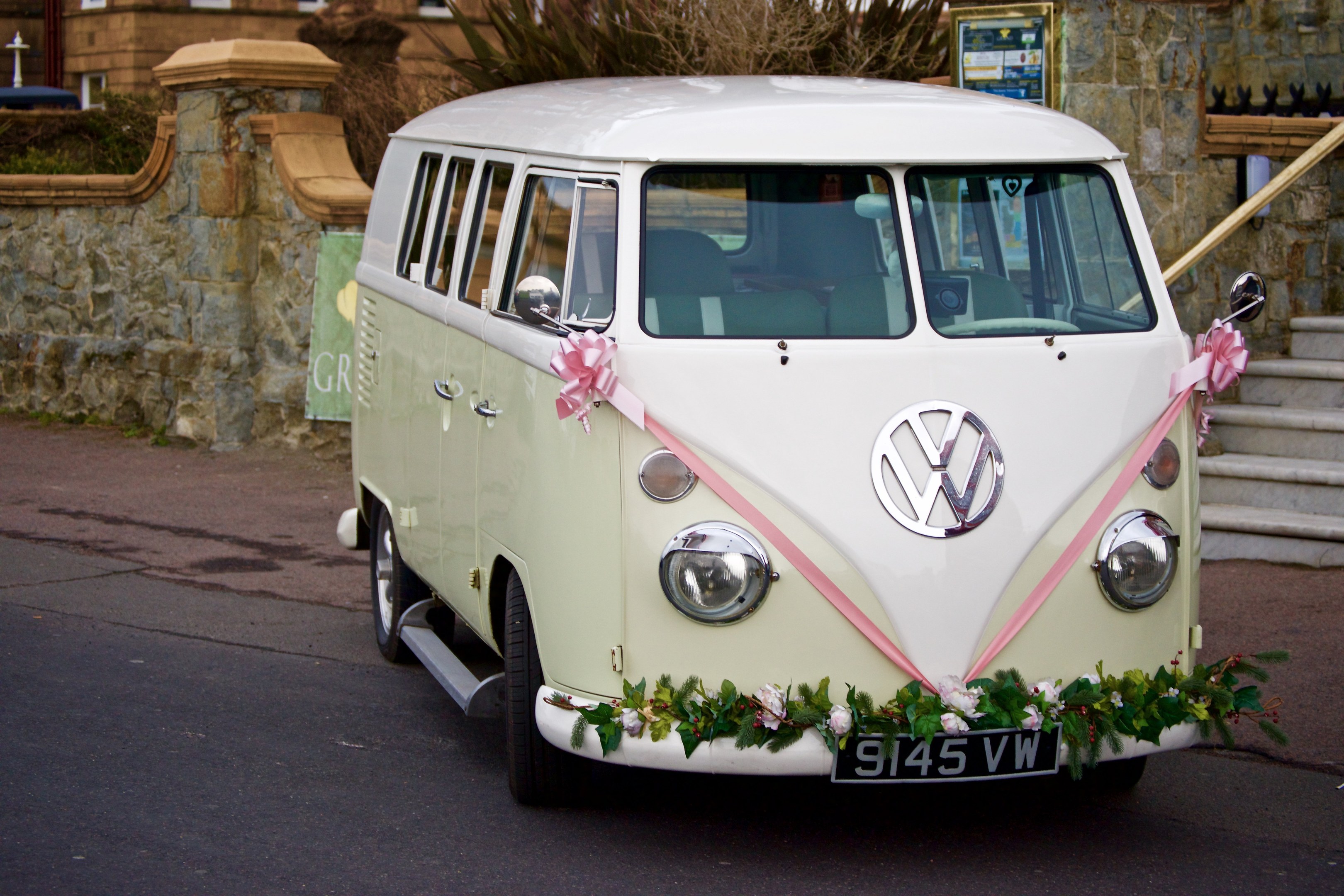 Ein wei├čer und rosafarbener VW Camper Van mit floralen Verzierungen, der vor einer Steinwand geparkt ist, mit einer Treppe auf der rechten Seite und einem Geb├Ąude im Hintergrund.