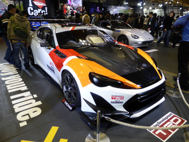 Eine Gruppe von Menschen um ein Rennfahrzeug auf der Tokyo Motor Show herumsteht, umgeben von Absperrpollern mit Seilen und Schildern mit Text und Bildern im Hintergrund unter beleuchteter Decke.