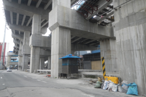 Große Betonstruktur im Bau mit darunterfahrenden Fahrzeugen, Baustoffe und ein Zelt auf der rechten Seite und Gebäude im Hintergrund.
