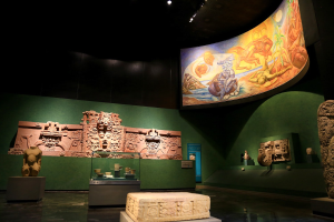 Eine Museumsausstellung im Anthropologischen Museum von Mexiko-Stadt, die verschiedene Artefakte zeigt, darunter Skulpturen, Gegenstände auf Tischen, eine Texttafel, eine Wandmalerei und Deckenleuchten.