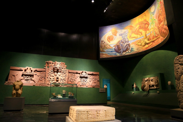 Eine Museumsausstellung im Anthropologischen Museum von Mexiko-Stadt, die verschiedene Artefakte zeigt, darunter Skulpturen, Gegenstände auf Tischen, eine Texttafel, eine Wandmalerei und Deckenleuchten.
