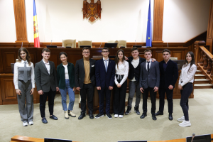 Eine Gruppe von Menschen, die Gewinner des Europäischen Gerichtshofs 2018-2019, lächeln und posieren für ein Foto vor einem Gerichtssaal mit Monitoren, Mikrofonen, einer hölzernen Plattform mit Stühlen, Flaggen und einem Objekt an der Wand, Treppen und Geländern.