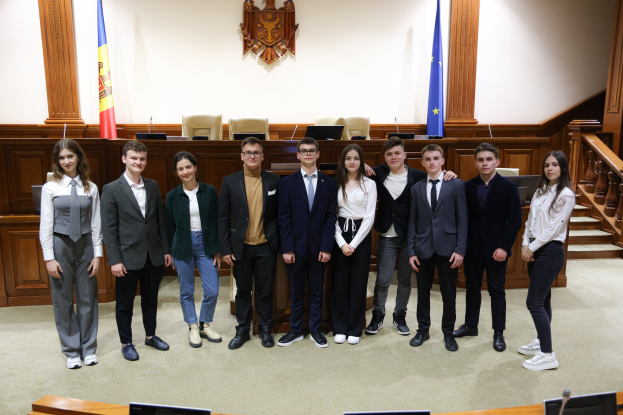 Eine Gruppe von Menschen, die Gewinner des Europäischen Gerichtshofs 2018-2019, lächeln und posieren für ein Foto vor einem Gerichtssaal mit Monitoren, Mikrofonen, einer hölzernen Plattform mit Stühlen, Flaggen und einem Objekt an der Wand, Treppen und Geländern.