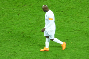 Ein Fußballspieler in einem weißen T-Shirt, weißen Shorts und orangefarbenen Schuhen läuft über ein grünes Feld.