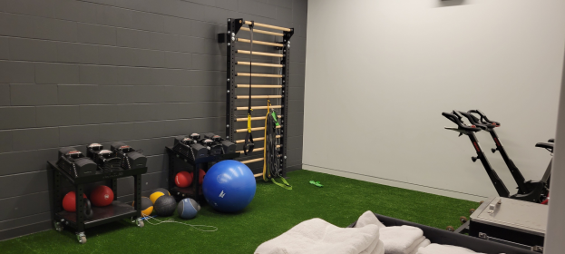 Ein Heimfitnessstudio mit Hanteln, Medizinbällen und anderem Equipment auf Gestellen, Handtücher auf dem Boden und eine Wand mit einem Objekt im Hintergrund, alles auf einem Kunstgrasboden.