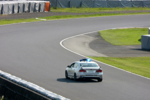 Ein Polizeiauto fährt auf einer Rennstrecke, mit Gras auf beiden Seiten und Menschen, Zelten und anderen Gegenständen im Hintergrund.