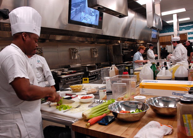 Eine Gruppe von Köchen in einer Küche beim Zubereiten von Lebensmitteln auf einem Schneidbrett, wobei ein Koch in der foreground in einer weißen Schürze und einer weißen Haube steht und ein Messer hält und von Utensilien, Schüsseln, Gemüse, einem Mobiltelefon, Flaschen und anderen Gegenständen umgeben ist, während im Hintergrund ein Fernseher an der Wand befestigt ist.