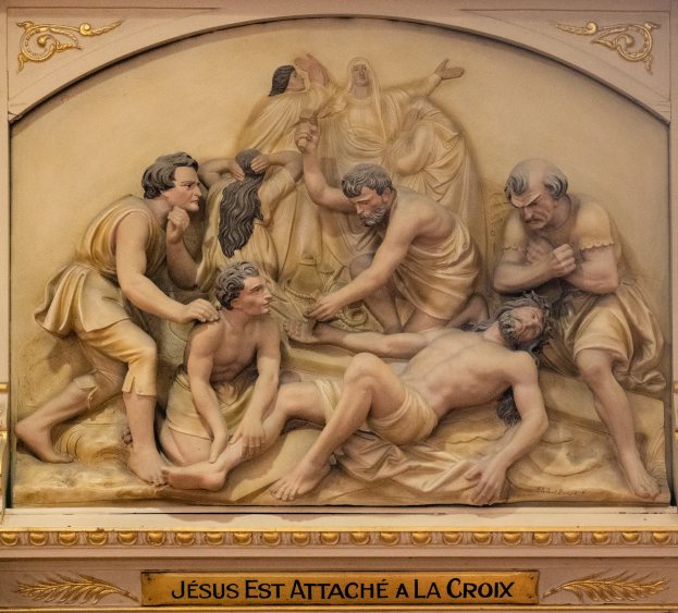 Eine Skulptur an einer Gebäudewand zeigt Jesus, der von einer Gruppe von Menschen angegriffen wird, mit einer Inschrift am Boden.