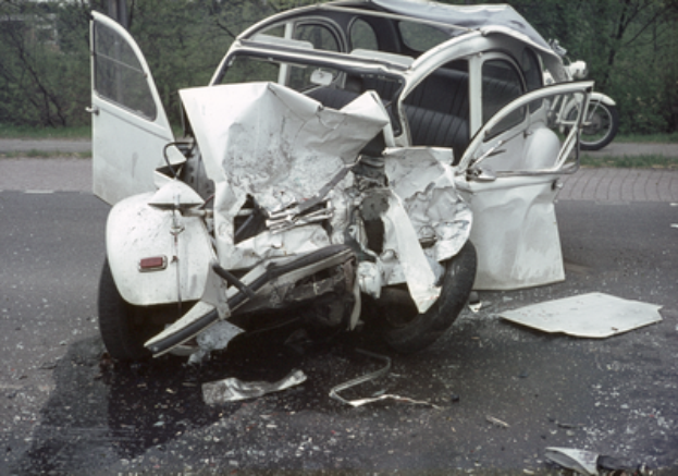 Ein stark vorne beschädigtes Auto steht am Straßenrand, mit Bäumen, einem Gebäude und einem anderen Fahrzeug im Hintergrund.