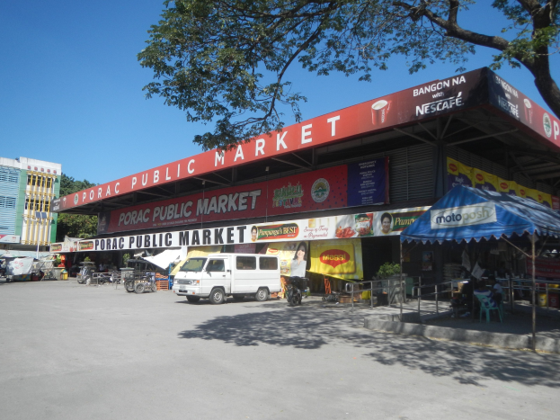 Ein öffentlicher Markt mit einem vor ihm geparkten Van, umgeben von Gebäuden, Geschäften, Schildern, Kraftfahrzeugen auf der Straße, Personen unter einem Zelt sitzend, Geländern, Bäumen und einem klaren blauen Himmel.